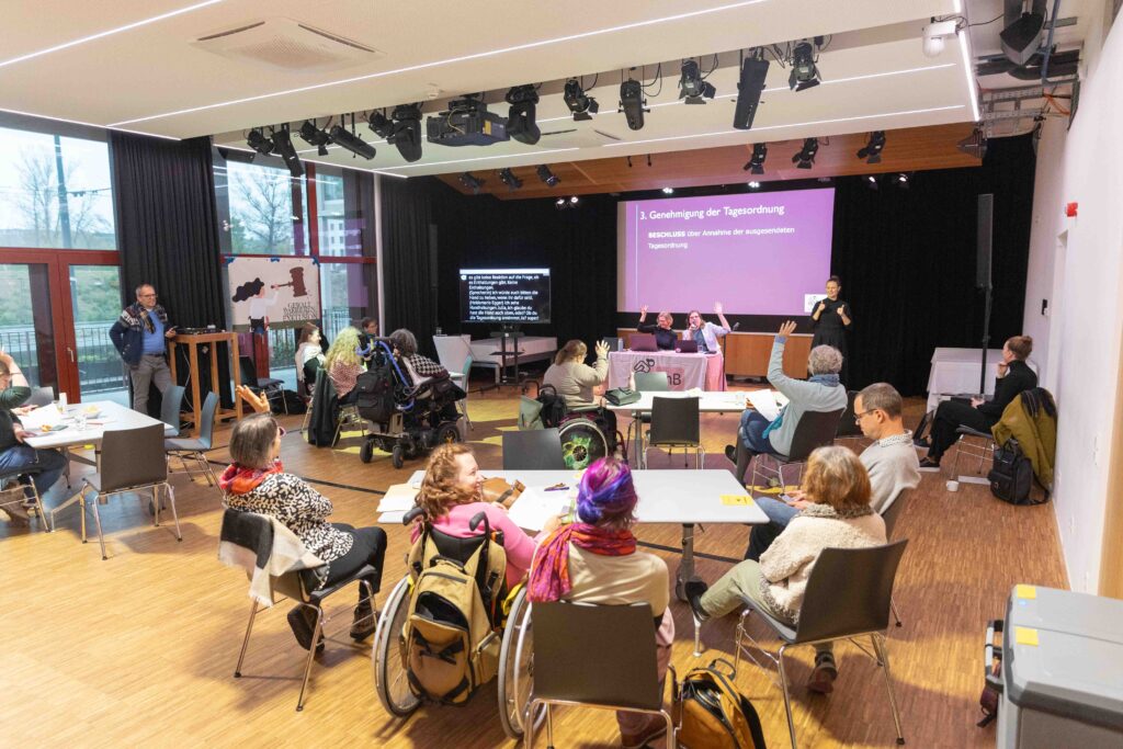 Ein Foto des gut gefüllten Nordbahnsaals. Frauen* mit Behinderungen sitzen um Tische herum verteilt, vorne bringen Julia und Heidemarie die Tagesordnung zur Abstimmung.