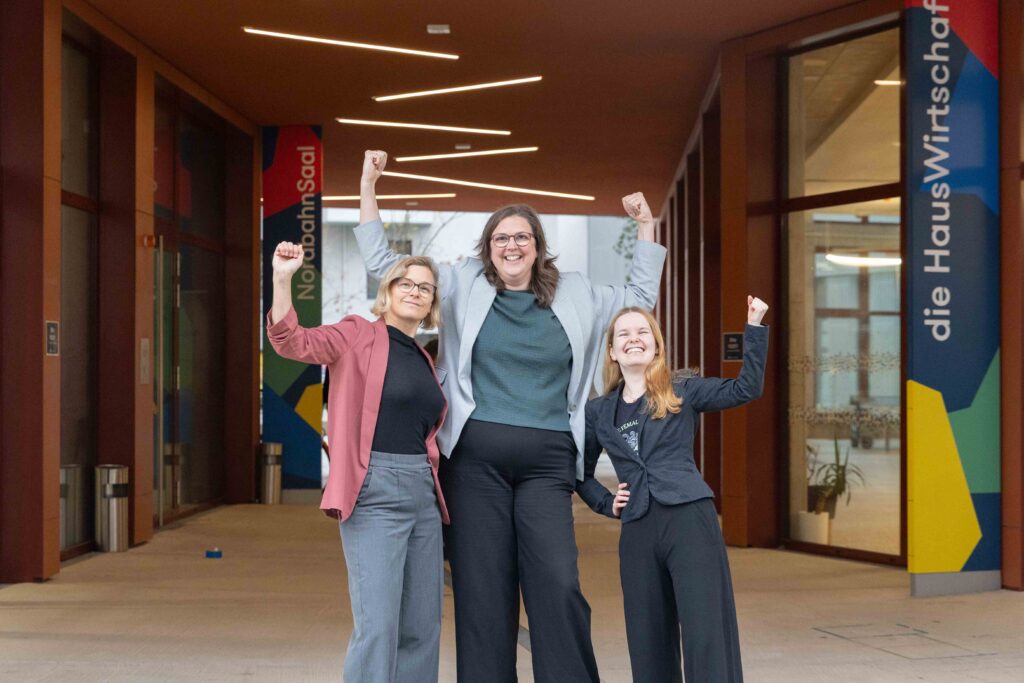 Ein Foto vom FmB-Team vor dem Nordbahnsaal. Julia, Heidi und Chrisi haben in einer stolzen Pose die Fäuste erhoben und lachen in die Kamera.