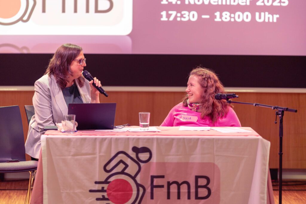 Ein Foto von Heidemarie (links) und Julia Hofmarcher (rechts), die gerade ihre Rede hielt. Heidemarie spricht ins Mikrofon, Julia lächelt sie an. Sie sitzen an einem Tisch, der mit dem FmB-Logo dekoriert ist.