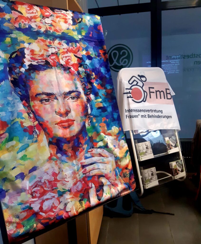 Ein farbenfrohes Gemälde mit einem Porträt von Frida Kahlo auf einer Staffelei. Daneben steht ein Prospektständer mit Flyern und einem Banner mit der Aufschrift „FmB – Interessenvertretung Frauen mit Behinderungen“. Im Hintergrund ist ein Fenster mit dem Logo von „Selbstbestimmt Leben“ zu sehen.
