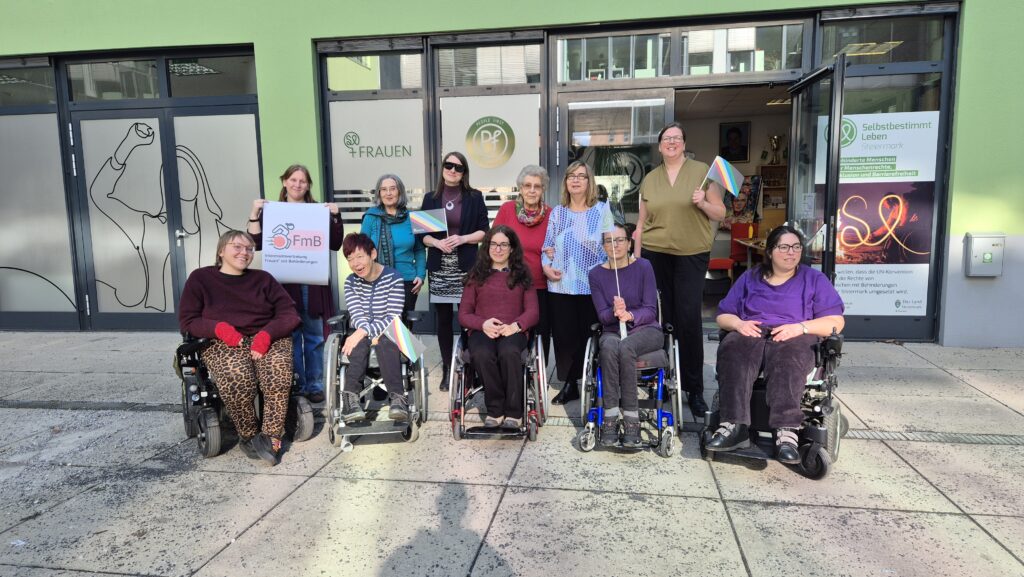 Gruppenfoto von elf Frauen vor dem Eingang eines Vereinsgebäudes. Einige der Frauen sitzen in Rollstühlen, andere stehen dahinter. Mehrere halten kleine Regenbogenfahnen in der Hand, eine Person hält ein Schild mit der Aufschrift „FmB – Interessenvertretung Frauen mit Behinderungen“. Im Hintergrund sind Glastüren mit Logos und Schriftzügen des Vereins „Selbstbestimmt Leben Steiermark“ zu sehen. Die Gruppe lächelt in die Kamera.
