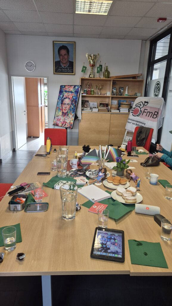 Ein heller Seminarraum mit einem großen Besprechungstisch. Auf dem Tisch stehen Wassergläser, Karaffen, Notizblöcke, Mobiltelefone und ein Tablet. In der Mitte liegen Informationsmaterialien, kleine Regenbogenfähnchen, eine Topfpflanze sowie Teller mit Krapfen. Im Hintergrund befinden sich ein Regal mit Pokal und Unterlagen, ein Porträt an der Wand und ein Banner mit der Aufschrift „FmB – Interessenvertretung Frauen mit Behinderungen“ sowie „Selbstbestimmt Leben Steiermark“.
