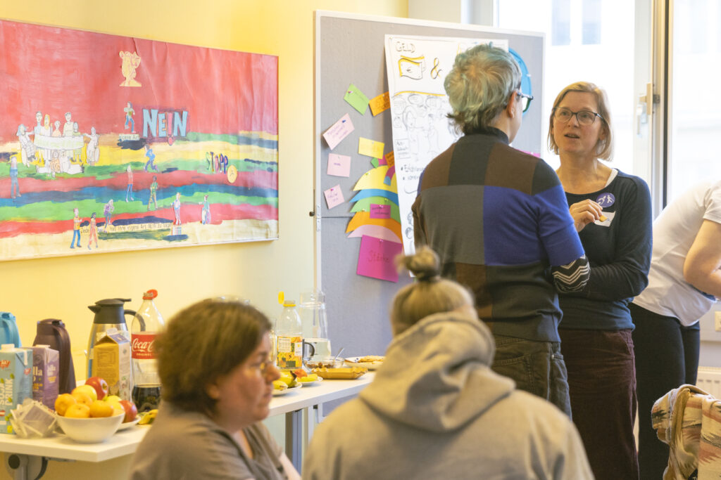 Austausch mit Buffet, stehende und sitzende Frauen mB sprechen miteinander, man sieht ien kleines Buffet