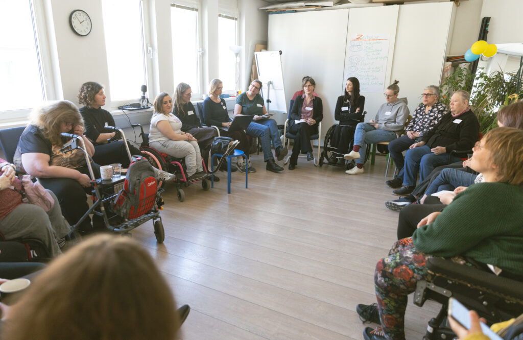 19 Frauen mit sichtbaren und unsichtbaren Behinderungen unterstützt von Frauen ohne Behinderungen sitzen im Kreis.