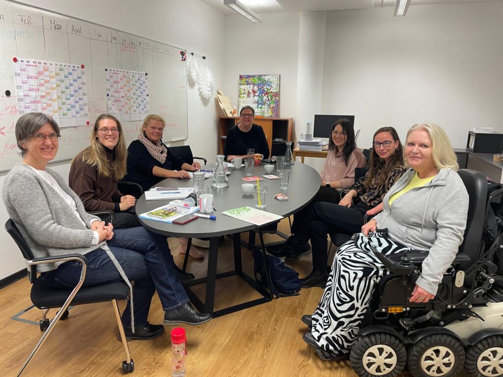 Sieben Frauen sitzen gemeinsam an einem runden Tisch in einem Besprechungsraum und lächeln in die Kamera. Auf dem Tisch stehen Gläser, Tassen, Notizen und Stifte. Im Hintergrund ist ein Whiteboard mit Kalenderübersicht und Notizen zu sehen. Eine Frau sitzt rechts im Bild in einem elektrischen Rollstuhl. Die Atmosphäre wirkt freundlich und kollegial.