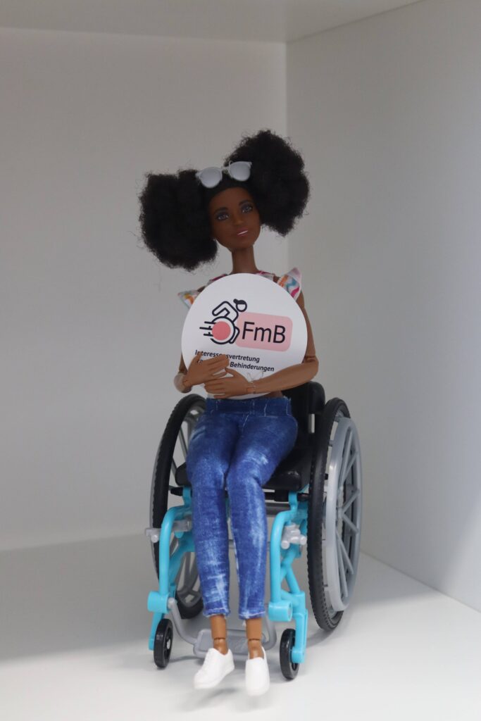 Eine schwarze Barbie-Puppe mit lockigem Haar sitzt in einem Rollstuhl. Sie trägt Jeans, weiße Schuhe und eine Sonnenbrille auf dem Kopf. In den Händen hält sie ein rundes Schild mit dem Logo „FmB – Interessensvertretung Frauen* mit Behinderungen“. Der Hintergrund ist schlicht und weiß.