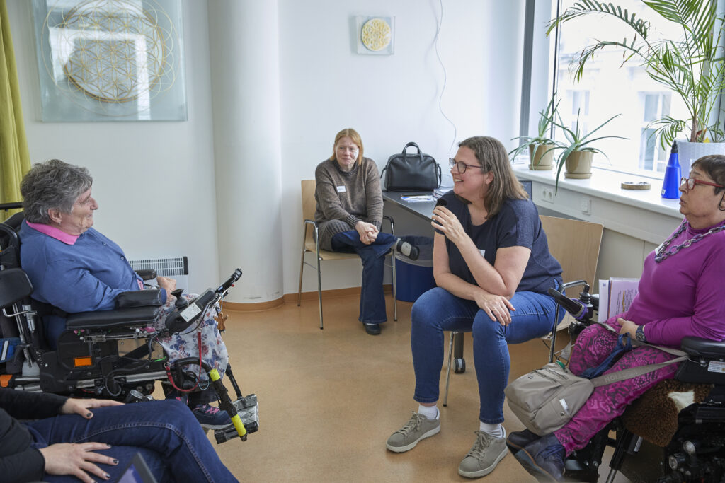 Eine kleine Gruppe von Erwachsenen sitzt in einem hellen Raum in einem Sesselkreis und führt ein Gespräch. Im Mittelpunkt spricht eine Frau mit einem Mikrofon und lächelt, während die anderen aufmerksam zuhören. Zwei Teilnehmerinnen* sitzen in elektrischen Rollstühlen. Im Hintergrund stehen Pflanzen auf der Fensterbank, und Tageslicht fällt durch große Fenster in den Raum. Die Atmosphäre wirkt offen, freundlich und respektvoll.