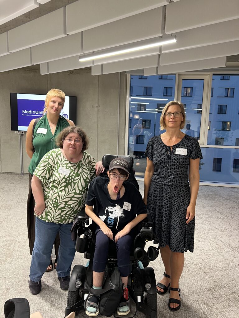 Vier Personen stehen für ein gemeinsames Foto bei einer MedInUnity-Veranstaltung zusammen. Zwei der abgebildeten Personen waren zuvor am Podium zu Gast, zwei weitere sind Mitglieder von FmB. Eine Person sitzt in einem elektrischen Rollstuhl, drei Personen stehen daneben. Im Hintergrund ist eine Präsentationsleinwand mit dem Schriftzug „MedInUnity“ zu sehen.