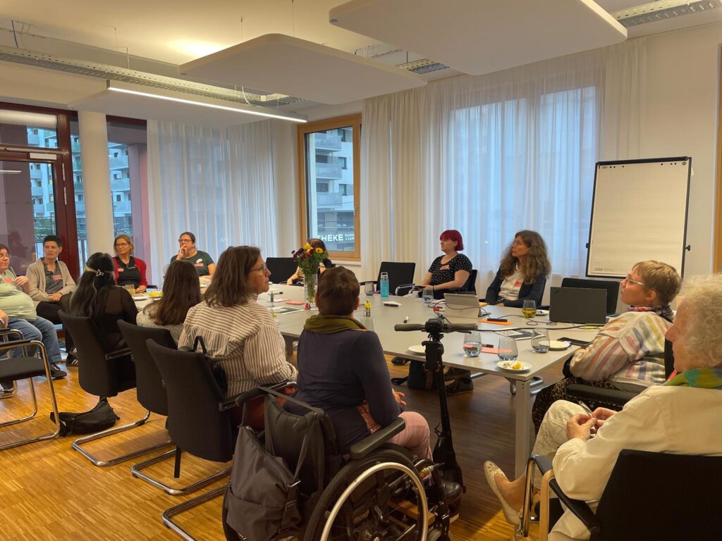 ca. 15 Frauen* mit unterschiedlichen Behinderungen, manche sichtbar und manche unsichtbar, sitzen um den langen Tisch herum und tauschen sich aus. 