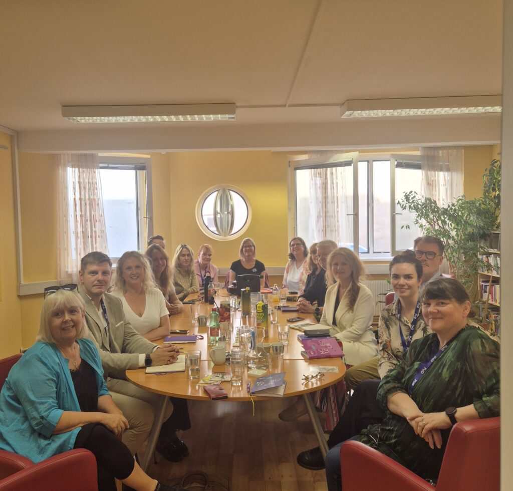 Julia und Heidi von FmB sitzen mit 13 Personen aus Island rund um einen Tisch. Alle schauen freundlich nach vorne in die Kamera.