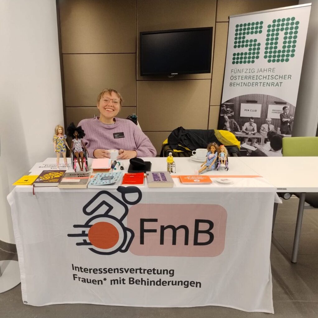 Ein Tisch mit einem weißen Tuch und dem Logo von FmB – Interessensvertretung Frauen* mit Behinderungen. Auf dem Tisch liegen Bücher, Broschüren und Informationsmaterialien; daneben stehen mehrere Puppen, darunter eine Puppe im Rollstuhl. Hinter dem Tisch sitzt eine lächelnde Person mit kurzen blonden Haaren und Brille.