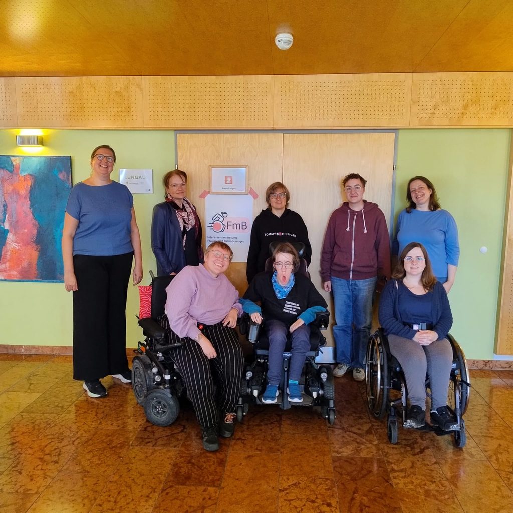 Acht Personen aus der FmB-Gruppe stehen oder sitzen für ein Gruppenfoto vor einem FmB-Schild in einem Raum im Lungau. Drei Personen sitzen in Rollstühlen, die anderen stehen dahinter oder daneben.