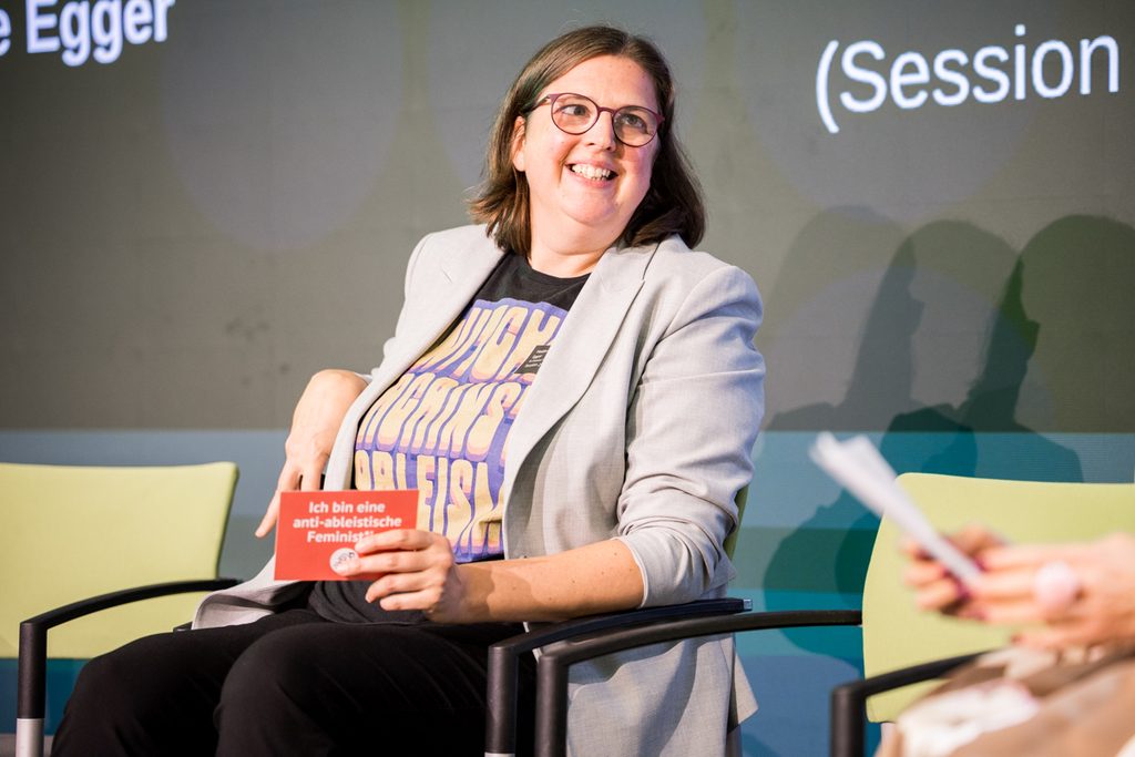Heidemarie Egger sitzt lächelnd auf einem Stuhl auf der Bühne der Fachkonferenz. Sie trägt eine helle Blazerjacke, eine Brille und ein T-Shirt mit einer feministischen Botschaft. In der Hand hält sie eine rote Karte mit der Aufschrift „Ich bin eine anti-ableistische Feministin“.
